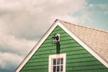 A green wooden house gable with white trim and a black outdoor lantern mounted above a window. The structure's shingles and the clear sky with clouds add texture and contrast to the scene.