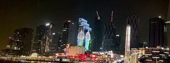 A nighttime cityscape featuring modern skyscrapers with bright lights and illuminated patterns. The buildings are tall and varied in architecture, including reflective surfaces and intricate designs. Vibrant light projections on a central building add a dynamic element to the scene. The ground level is bustling with smaller structures and lively lighting.