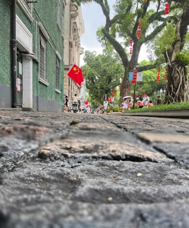 A cobblestone street lined with greenery and large trees featuring red lanterns and Chinese flags. People can be seen walking and riding bikes. The architecture includes Western-style buildings with green and beige facades, creating a blend of natural and structured elements.