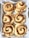 Six freshly baked cinnamon rolls placed on a sheet of parchment paper inside a baking tray. The rolls have a golden-brown crust with visible swirls of cinnamon. Each roll is neatly arranged, showcasing their spiral shape and cinnamon-coated tops.