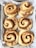 Six freshly baked cinnamon rolls placed on a sheet of parchment paper inside a baking tray. The rolls have a golden-brown crust with visible swirls of cinnamon. Each roll is neatly arranged, showcasing their spiral shape and cinnamon-coated tops.