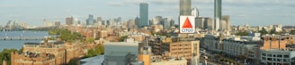 A panoramic cityscape featuring a mix of modern skyscrapers and historic brick buildings, with a prominent CITGO sign in the foreground. The skyline includes a variety of architectural styles, and a river can be seen alongside the city, with trees lining its banks.