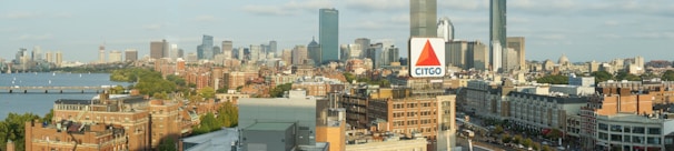 A panoramic cityscape featuring a mix of modern skyscrapers and historic brick buildings, with a prominent CITGO sign in the foreground. The skyline includes a variety of architectural styles, and a river can be seen alongside the city, with trees lining its banks.