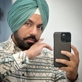 A man wearing a turquoise turban and patterned shirt is taking a selfie with a smartphone. He has a serious expression and is holding the phone in his right hand while his left hand is near his face.