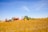 A green tractor is pulling a large agricultural implement across an open field, which is covered in golden stubble. The sky above is clear blue, and a few leafless trees are visible in the background, suggesting a rural farming scene.