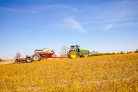 A green tractor is pulling a large agricultural implement across an open field, which is covered in golden stubble. The sky above is clear blue, and a few leafless trees are visible in the background, suggesting a rural farming scene.