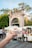 A hand is holding a white cup filled with a dessert topped with red sauce and shredded cheese or coconut. The background features a busy outdoor market with people and various stands. There is a large stone archway and trees in the distance.