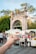 A hand is holding a white cup filled with a dessert topped with red sauce and shredded cheese or coconut. The background features a busy outdoor market with people and various stands. There is a large stone archway and trees in the distance.