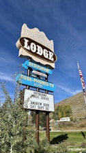 A vintage-style sign advertises The Virginian Lodge with multiple segments displaying offerings such as a restaurant and a music series. The sign includes details for an event featuring Andrew Bird on September 30. It is set against a backdrop of a bright blue sky and a grassy landscape with trees and an American flag visible.