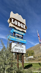 A vintage-style sign advertises The Virginian Lodge with multiple segments displaying offerings such as a restaurant and a music series. The sign includes details for an event featuring Andrew Bird on September 30. It is set against a backdrop of a bright blue sky and a grassy landscape with trees and an American flag visible.