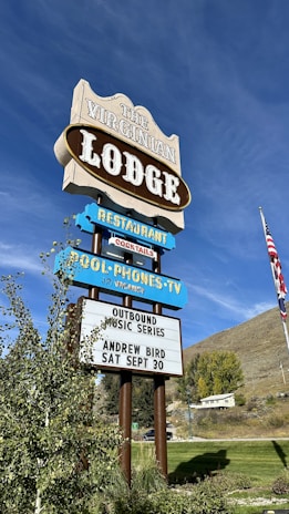 A vintage-style sign advertises The Virginian Lodge with multiple segments displaying offerings such as a restaurant and a music series. The sign includes details for an event featuring Andrew Bird on September 30. It is set against a backdrop of a bright blue sky and a grassy landscape with trees and an American flag visible.