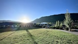 A landscape features a sunlit grassy area with long shadows stretching across a well-maintained lawn. In the background, a range of green hills or mountains can be seen under a clear blue sky. A structure, possibly a pavilion or gazebo, is situated near manicured gardens with various trees and plants.