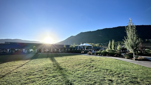 A landscape features a sunlit grassy area with long shadows stretching across a well-maintained lawn. In the background, a range of green hills or mountains can be seen under a clear blue sky. A structure, possibly a pavilion or gazebo, is situated near manicured gardens with various trees and plants.