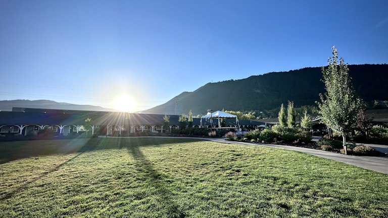 A landscape features a sunlit grassy area with long shadows stretching across a well-maintained lawn. In the background, a range of green hills or mountains can be seen under a clear blue sky. A structure, possibly a pavilion or gazebo, is situated near manicured gardens with various trees and plants.