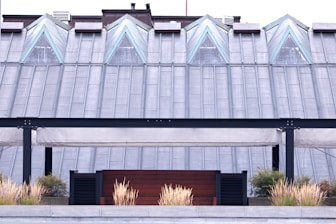 A modern rooftop structure featuring triangular glass windows positioned symmetrically along a metal-clad roof. Below, a patio area with beige, canopy-like awnings and black metal framing is fronted by decorative grass plants in a planter.