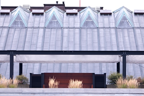 A modern rooftop structure featuring triangular glass windows positioned symmetrically along a metal-clad roof. Below, a patio area with beige, canopy-like awnings and black metal framing is fronted by decorative grass plants in a planter.
