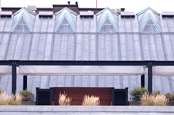 A modern rooftop structure featuring triangular glass windows positioned symmetrically along a metal-clad roof. Below, a patio area with beige, canopy-like awnings and black metal framing is fronted by decorative grass plants in a planter.