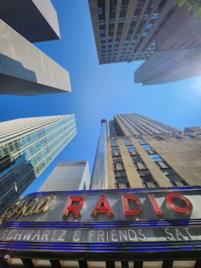 Tall skyscrapers stretch upward towards a clear blue sky, framing the view. In the foreground, a vintage-style marquee sign displays illuminated red letters that spell 'RADIO'. The buildings exhibit modern architectural styles with glass and stone exteriors.