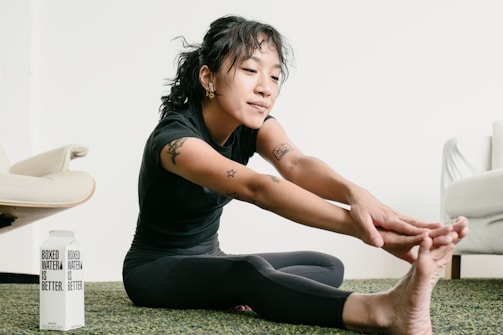 A person is performing a seated forward fold stretch on a carpeted floor, reaching toward their toes. They are wearing a black outfit and have tattoos on their arms. Next to them is a carton labeled 'Boxed Water Is Better'. In the background, there's a light-colored chair and sofa.