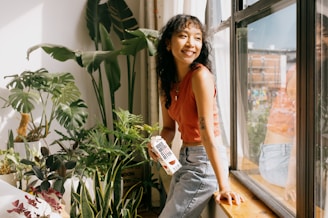 A person with curly hair and an orange sleeveless top sits on a wooden windowsill, surrounded by a variety of green plants. They hold a carton labeled 'Boxed Water Is Better,' and sunlight streams through the large window, casting bright reflections and creating a serene indoor gardening atmosphere.