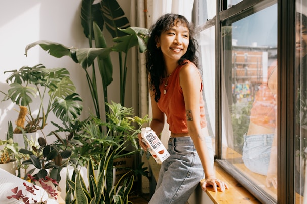 A person with curly hair and an orange sleeveless top sits on a wooden windowsill, surrounded by a variety of green plants. They hold a carton labeled 'Boxed Water Is Better,' and sunlight streams through the large window, casting bright reflections and creating a serene indoor gardening atmosphere.