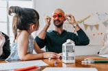 A child and an adult are sitting at a table scattered with colorful pencils and paper. The adult is wearing playful, oversized orange glasses and engaging with the child. A carton labeled 'Boxed Water Is Better' is prominently placed on the table.