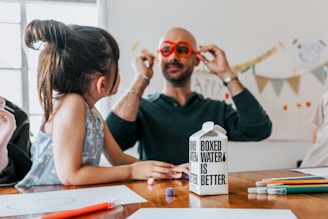A child and an adult are sitting at a table scattered with colorful pencils and paper. The adult is wearing playful, oversized orange glasses and engaging with the child. A carton labeled 'Boxed Water Is Better' is prominently placed on the table.