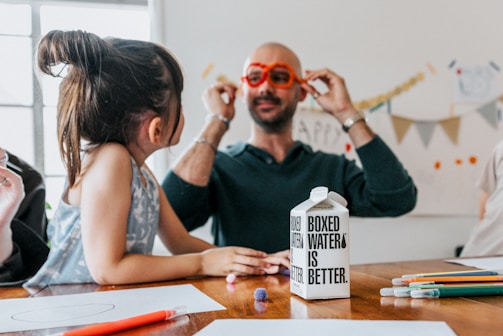 A child and an adult are sitting at a table scattered with colorful pencils and paper. The adult is wearing playful, oversized orange glasses and engaging with the child. A carton labeled 'Boxed Water Is Better' is prominently placed on the table.