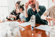 A family is sitting at a wooden table engaged in a crafting activity. An adult with red glasses is handling some boxes labeled 'boxed water is better'. Two children and another adult are also present, using colored pens and small colorful craft balls. The scene is brightly lit, suggesting a casual and relaxed atmosphere.