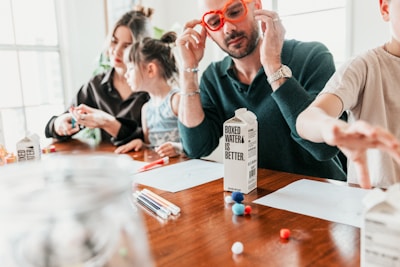 A family is sitting at a wooden table engaged in a crafting activity. An adult with red glasses is handling some boxes labeled 'boxed water is better'. Two children and another adult are also present, using colored pens and small colorful craft balls. The scene is brightly lit, suggesting a casual and relaxed atmosphere.