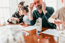 A family is sitting at a wooden table engaged in a crafting activity. An adult with red glasses is handling some boxes labeled 'boxed water is better'. Two children and another adult are also present, using colored pens and small colorful craft balls. The scene is brightly lit, suggesting a casual and relaxed atmosphere.