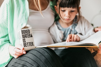 A person in a green-striped shirt holds a carton with 'Boxed Water Is Better' printed on it. Beside them, a child is focused on reading a book. The atmosphere is calm and intimate, suggesting a moment of learning or bonding.