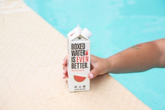 A hand holding a carton labeled 'Boxed Water Is Even Better' with watermelon branding. The hand is resting on a textured surface beside a swimming pool.