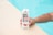 A hand holding a carton labeled 'Boxed Water Is Even Better' with watermelon branding. The hand is resting on a textured surface beside a swimming pool.