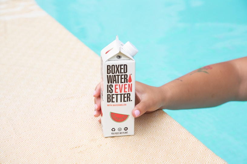 A hand holding a carton labeled 'Boxed Water Is Even Better' with watermelon branding. The hand is resting on a textured surface beside a swimming pool.