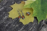 A pair of silver earrings and a matching ring with amber-colored stones placed on top of a large yellow maple leaf, with a green maple leaf partially overlapping. The leaves rest on a rough, weathered wooden surface.