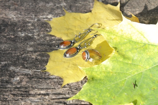 A pair of silver earrings and a ring with orange stones rest atop large yellow autumn leaves, placed on a rustic wooden surface.
