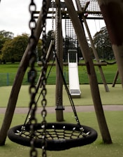An outdoor playground with a focus on a large net swing and a slide in the background. The playground equipment is made of wood and metal chains, set on a green rubberized surface with grassy fields and trees visible in the distance.