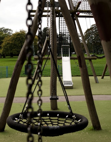 An outdoor playground with a focus on a large net swing and a slide in the background. The playground equipment is made of wood and metal chains, set on a green rubberized surface with grassy fields and trees visible in the distance.