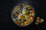 A smoothie bowl topped with a variety of fruits and nuts. Visible ingredients include sliced bananas, blueberries, mango chunks, chia seeds, pistachios, and what appears to be dried apricot. The bowl is arranged in a visually pleasing manner and surrounded by a dark surface, with a few physalis fruits placed outside the bowl.