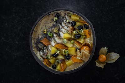 A smoothie bowl topped with a variety of fruits and nuts. Visible ingredients include sliced bananas, blueberries, mango chunks, chia seeds, pistachios, and what appears to be dried apricot. The bowl is arranged in a visually pleasing manner and surrounded by a dark surface, with a few physalis fruits placed outside the bowl.