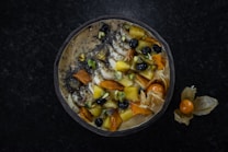 A smoothie bowl topped with a variety of fruits and nuts. Visible ingredients include sliced bananas, blueberries, mango chunks, chia seeds, pistachios, and what appears to be dried apricot. The bowl is arranged in a visually pleasing manner and surrounded by a dark surface, with a few physalis fruits placed outside the bowl.