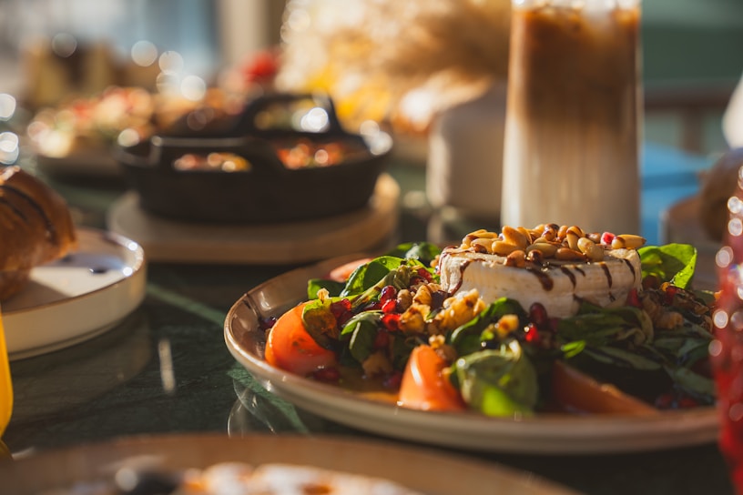 A fresh salad with vibrant greens, sliced tomatoes, walnuts, pomegranate seeds, and a wheel of cheese topped with pine nuts. The dish is set in a warm, sunlit atmosphere giving it an inviting and appetizing look. In the background, a glass of iced coffee stands tall, accompanied by a croissant and other food items.