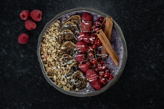 A beautifully arranged smoothie bowl with layers of granola, sliced figs, fresh raspberries, red currants, and two cinnamon sticks on top. The smoothie base appears to be a deep purple color, possibly acai or berry-flavored. A drizzle of white yogurt or cream decorates the top. Three raspberries are placed on the dark marble surface nearby.