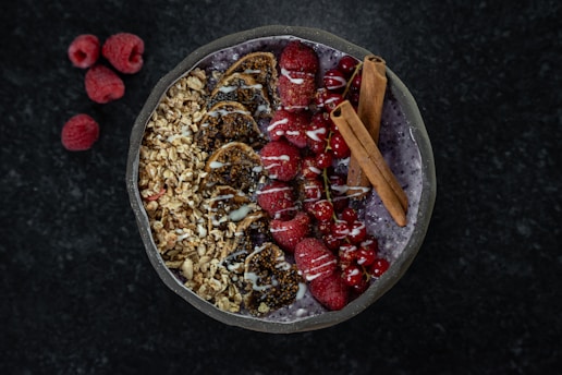 A beautifully arranged smoothie bowl with layers of granola, sliced figs, fresh raspberries, red currants, and two cinnamon sticks on top. The smoothie base appears to be a deep purple color, possibly acai or berry-flavored. A drizzle of white yogurt or cream decorates the top. Three raspberries are placed on the dark marble surface nearby.