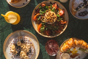 A spread of various breakfast or brunch dishes on a green marble table. There is a plate with a salad consisting of spinach, tomatoes, pine nuts, and a round piece of cheese topped with more nuts. Another dish features a waffle topped with banana slices, blueberries, and a sprinkle of powdered sugar. A croissant is also present, stuffed with cream and garnished with berries. Accompanying the meal are drinks, including a glass of orange juice with a yellow straw and a glass of red beverage.