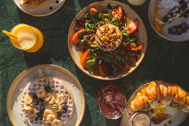 A spread of various breakfast or brunch dishes on a green marble table. There is a plate with a salad consisting of spinach, tomatoes, pine nuts, and a round piece of cheese topped with more nuts. Another dish features a waffle topped with banana slices, blueberries, and a sprinkle of powdered sugar. A croissant is also present, stuffed with cream and garnished with berries. Accompanying the meal are drinks, including a glass of orange juice with a yellow straw and a glass of red beverage.