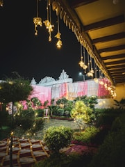 An outdoor setting decorated with colorful drapes in pink and white, with hanging lights creating a festive and inviting atmosphere. A single chair is placed among lush greenery and well-manicured plants, enhancing the garden-like environment. The setting is illuminated by warm, glowing lights, highlighting the intricate patterns on the ground covering.