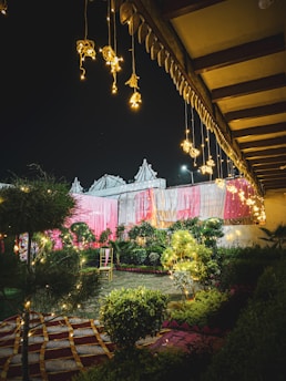 An outdoor setting decorated with colorful drapes in pink and white, with hanging lights creating a festive and inviting atmosphere. A single chair is placed among lush greenery and well-manicured plants, enhancing the garden-like environment. The setting is illuminated by warm, glowing lights, highlighting the intricate patterns on the ground covering.