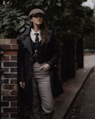 A person wearing a vintage-inspired outfit stands against a brick wall. The attire includes a newsboy cap, a tie, a waistcoat, and a long coat. The background is lush with greenery, giving a moody and classic feel.